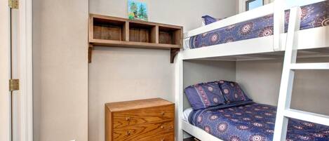 Uma cama de beliche com roupa de cama estampada, um penteou de madeira e uma estante com uma pequena pintura.