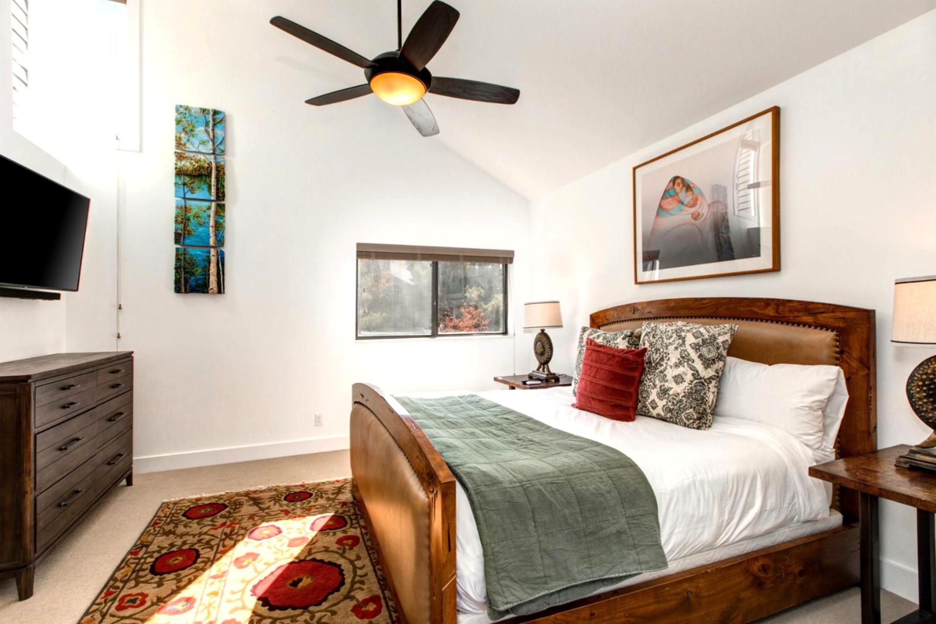 A bedroom with a wooden bed, a ceiling fan, a window, a TV, and artwork on the walls.