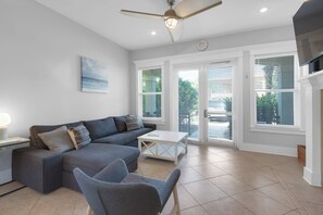 A modern living room with a grey sectional sofa, a white coffee table, and a grey armchair. There is a ceiling fan, a painting on the wall, and large windows with blinds.