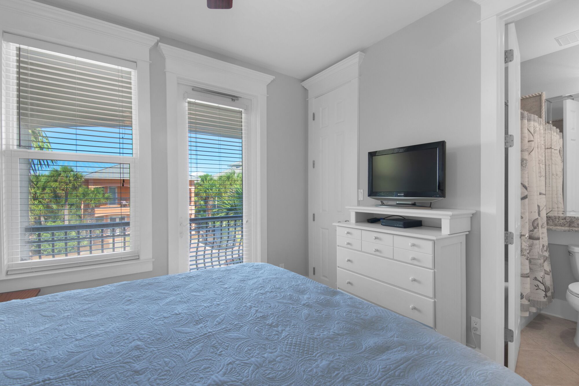 Un dormitorio con cama, televisor en una cómoda blanca y balcón con persianas.