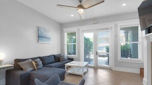 A modern living room with a grey sectional sofa, a white coffee table, and a grey armchair. There is a ceiling fan, a painting on the wall, and large windows with blinds. - Townhome-Comm Pool-Private Beach Access 5 Min Walk (Miramar Beach)
