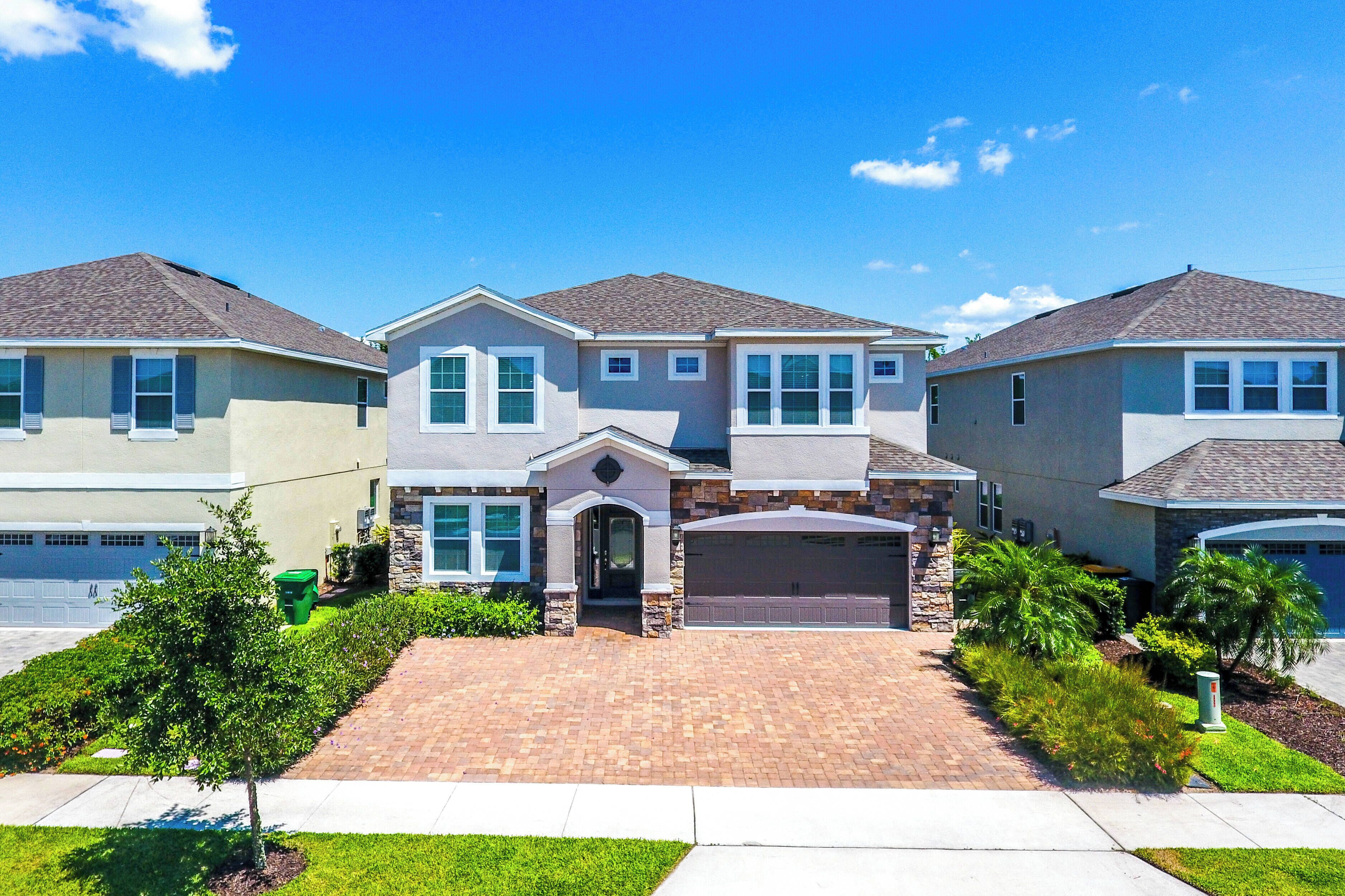 A suburban three-story house with a brick driveway, a garage, and a well-maintained lawn.