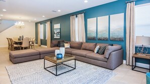 A modern living room with a sectional sofa, a glass coffee table, and a dining area with a table and chairs. - Gorgeous Single Home With Private Pool at Encore (Kissimmee)