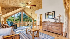 A spacious room with a wooden ceiling, a large window, a wooden bench, a wooden dresser, and a ceiling fan. - Big Bear Lodge Retreat where friends & families gather and meet! (Comins)