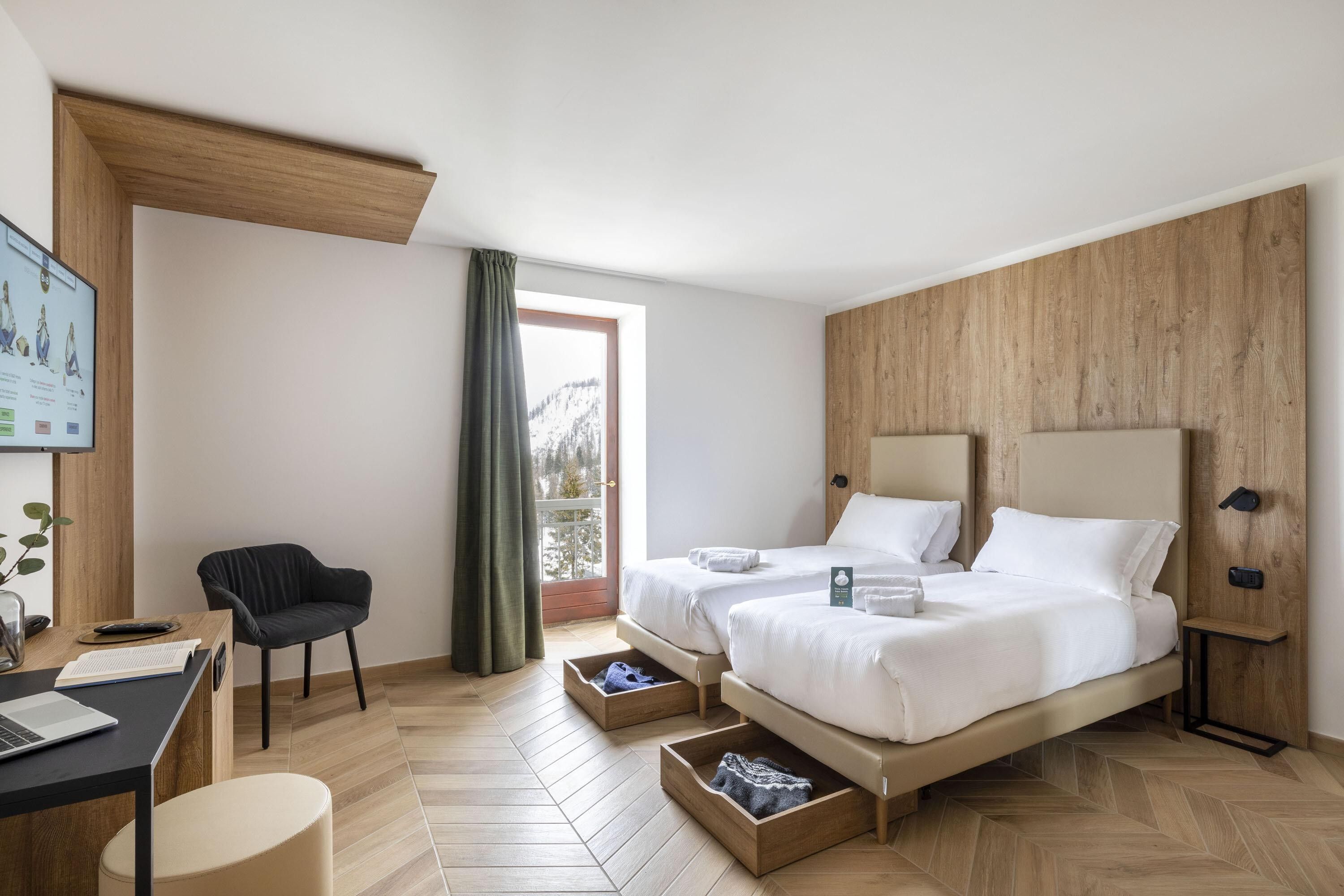 Una habitación de hotel moderna con piso de madera, un escritorio con una laptop, una silla, dos camas con ropa blanca y un ventanal con vistas a montañas nevadas.