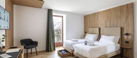 Una habitación de hotel moderna con piso de madera, un escritorio con una laptop, una silla, dos camas con ropa blanca y un ventanal con vistas a montañas nevadas.