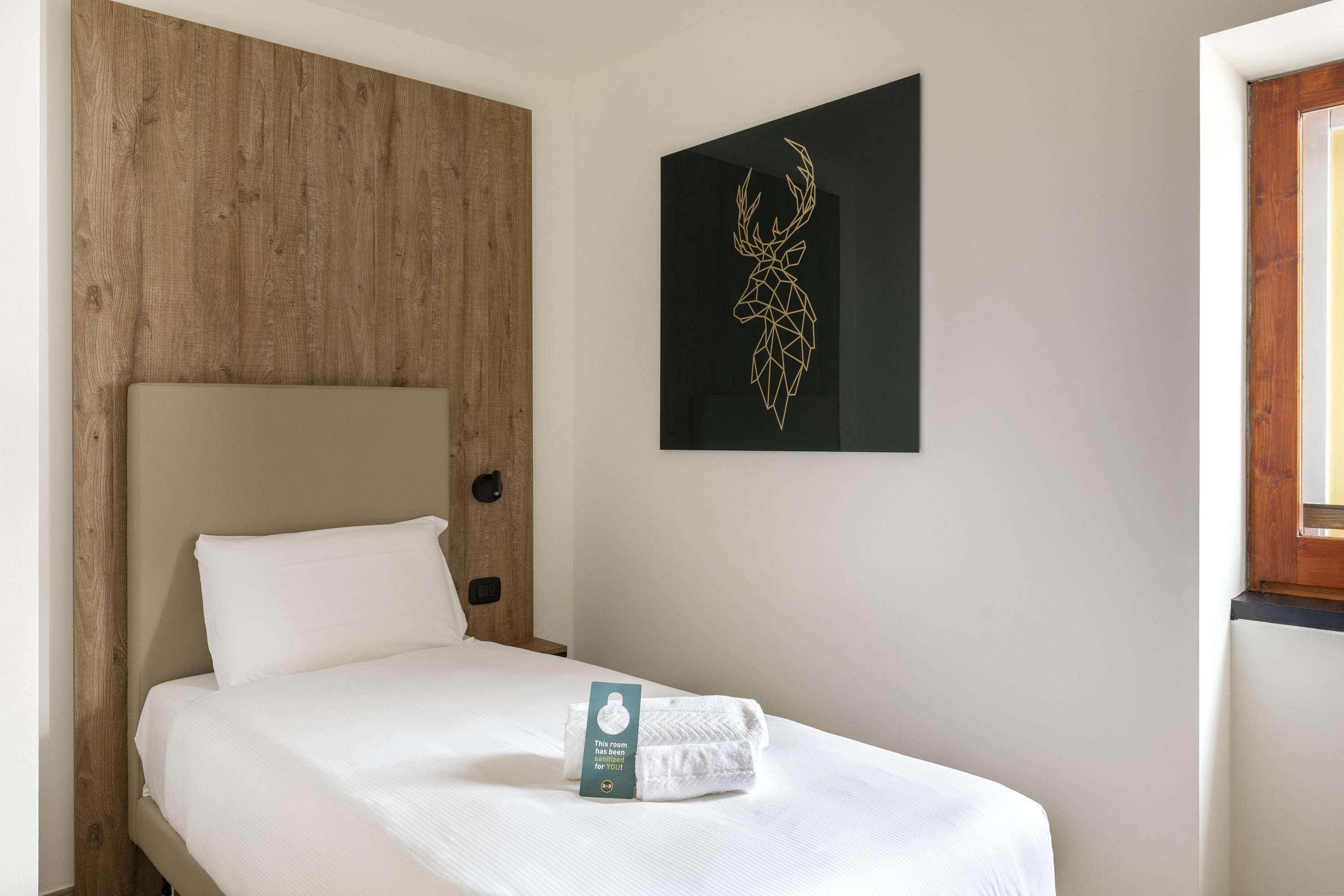 A hotel room with a wooden headboard, a bed with white bedding, a framed deer head artwork on the wall, and a window with a wooden frame.