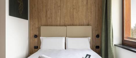 A neatly made bed with two pillows, a wooden headboard, and a window with curtains.