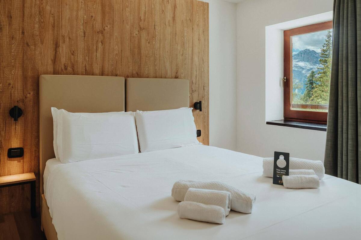 A bed with white linens, a wooden headboard, and a window with a mountain view.