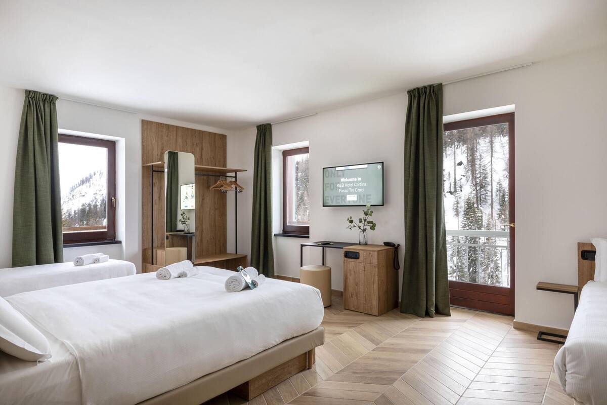 A hotel room with two beds, a desk, a TV, and a view of trees.