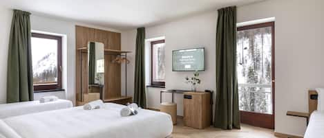A hotel room with two beds, a desk, a TV, and a view of trees.