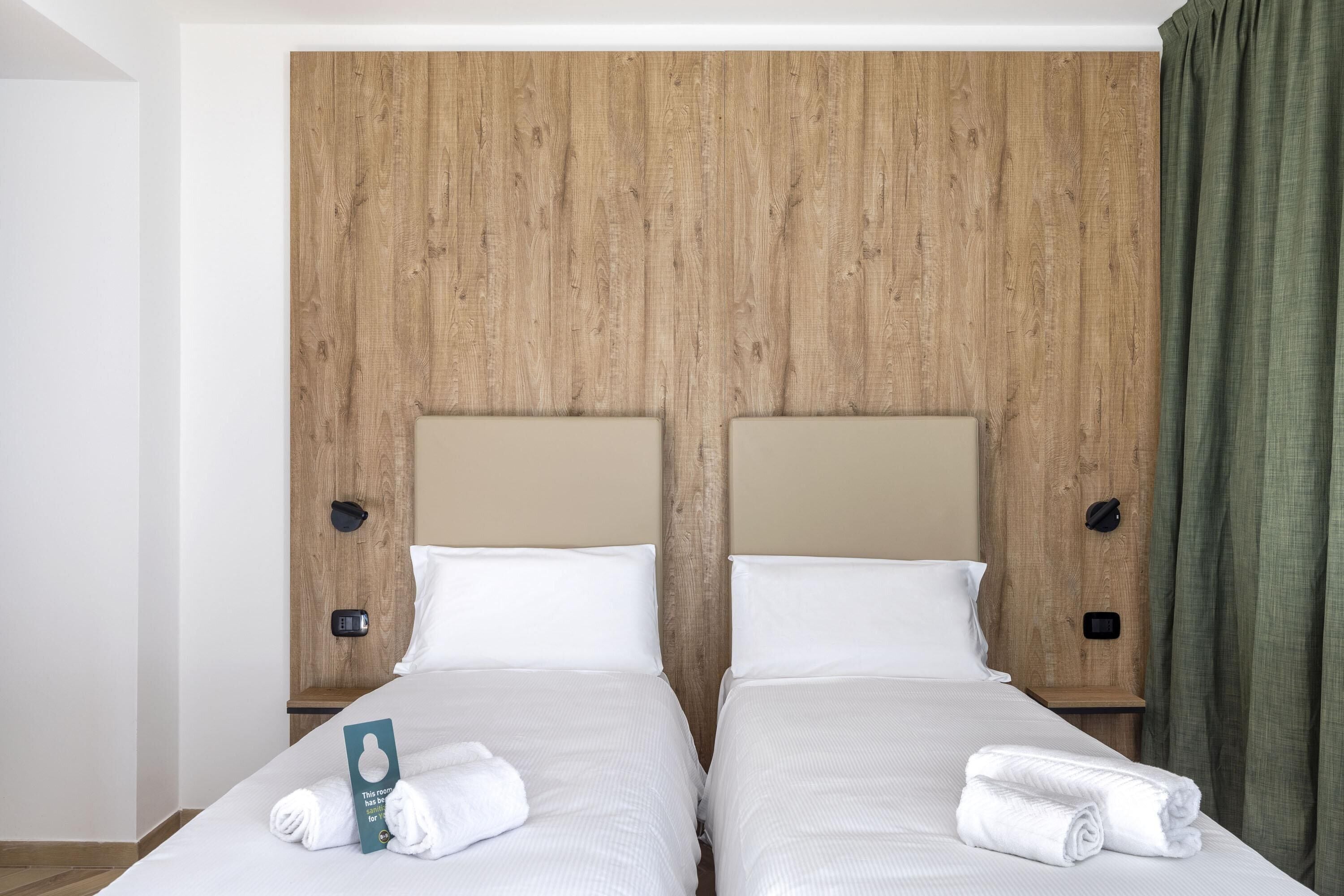 Un lit double avec du linge de lit blanc, deux tables de chevet, une tête de lit en bois et un rideau vert.