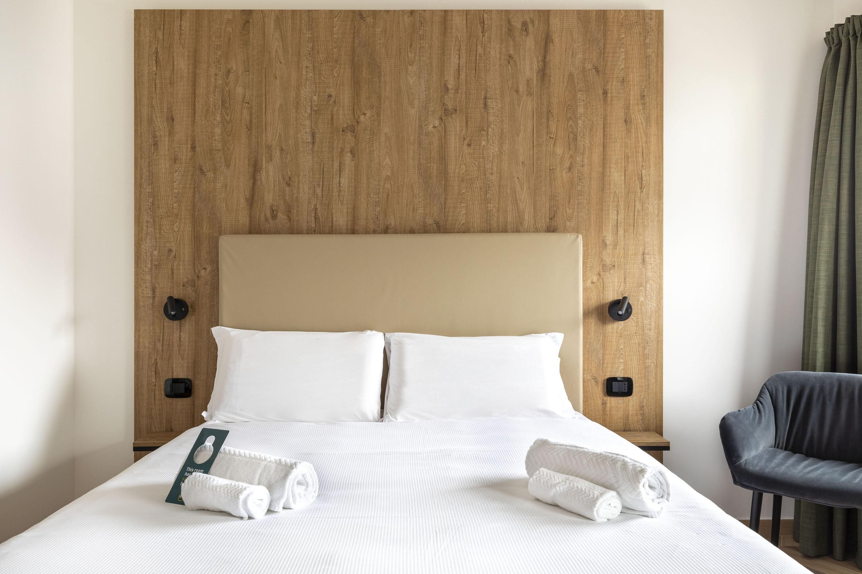 Une chambre d’hôtel avec une tête de lit en bois, du linge de lit blanc, une chaise noire et une paire de serviettes sur le lit.