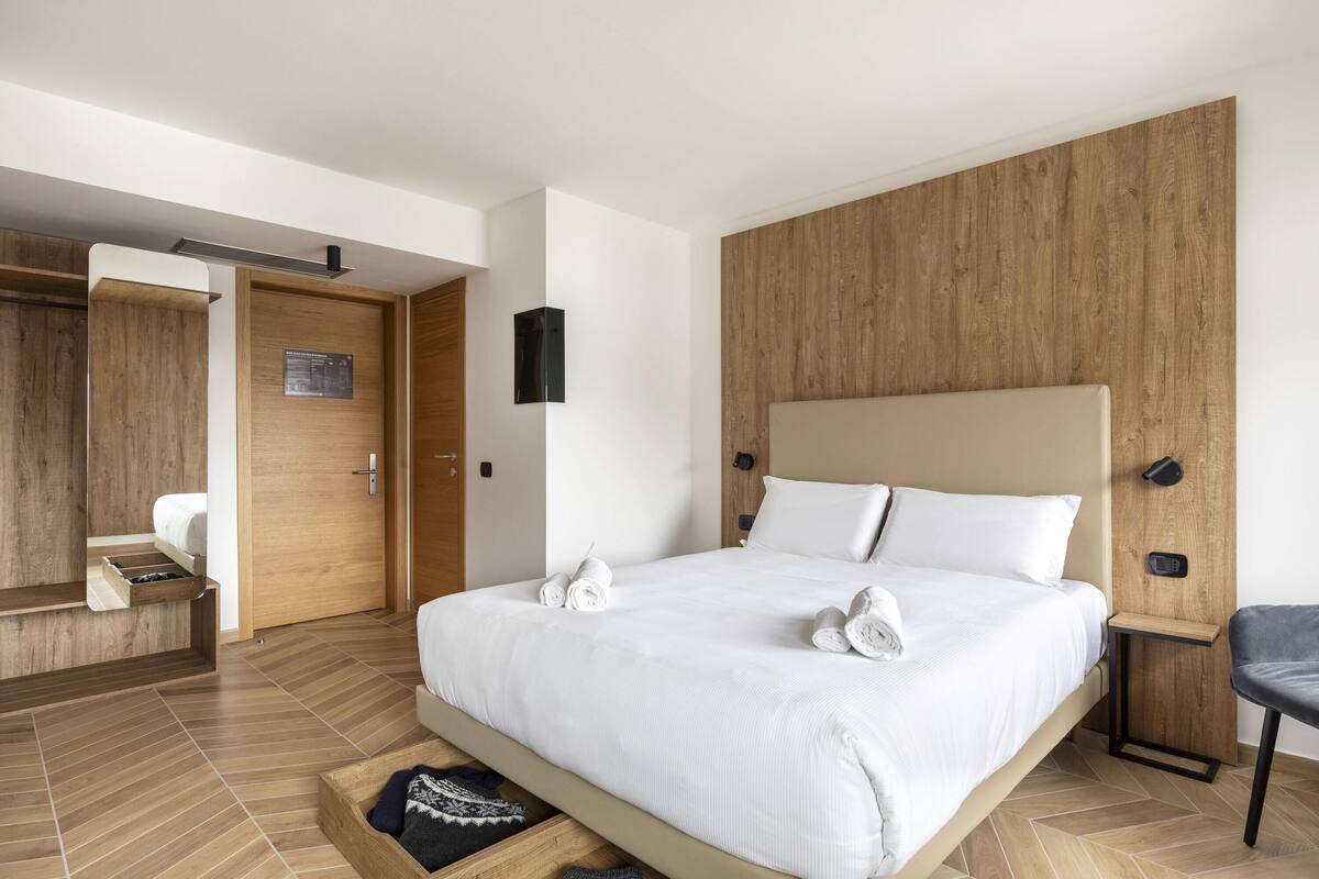 A modern hotel room with a large bed, wooden paneling on the walls, a built-in wardrobe, and a small seating area.