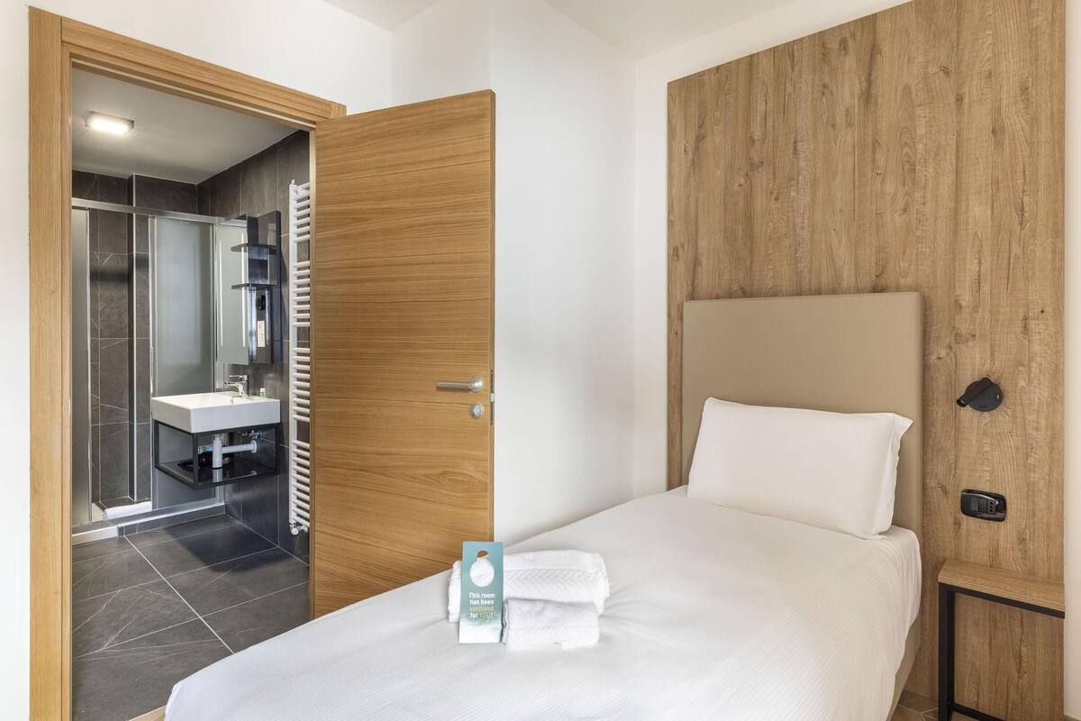 A hotel room with a bed, a wooden headboard, a bedside table, a nightstand, a wall-mounted light, and a view of the bathroom.