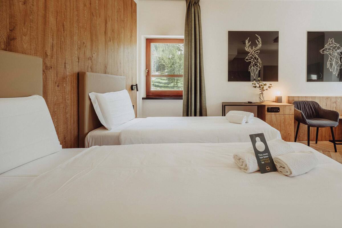 A hotel room with two beds, a desk, a chair, a window with a view of greenery, and artwork on the wall.