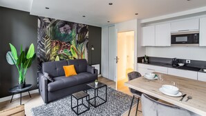 A modern living room with a dark sofa, a coffee table, and a dining area with a wooden table and chairs. - Le Kipling - Studio de Standing à 5 min du centre (Rennes)