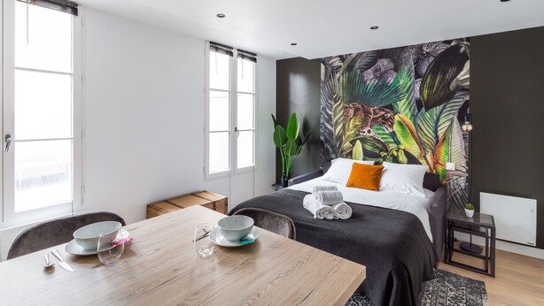 A modern hotel room with a large bed, a dining table with a bowl and glasses, and a tropical-themed wall mural. - Le Kipling - Studio de Standing à 5 min du centre (Rennes)