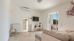 Un grand salon avec un canapé beige, un bureau blanc et un téléviseur. On y trouve également un radiateur et une fenêtre donnant sur un paysage verdoyant.