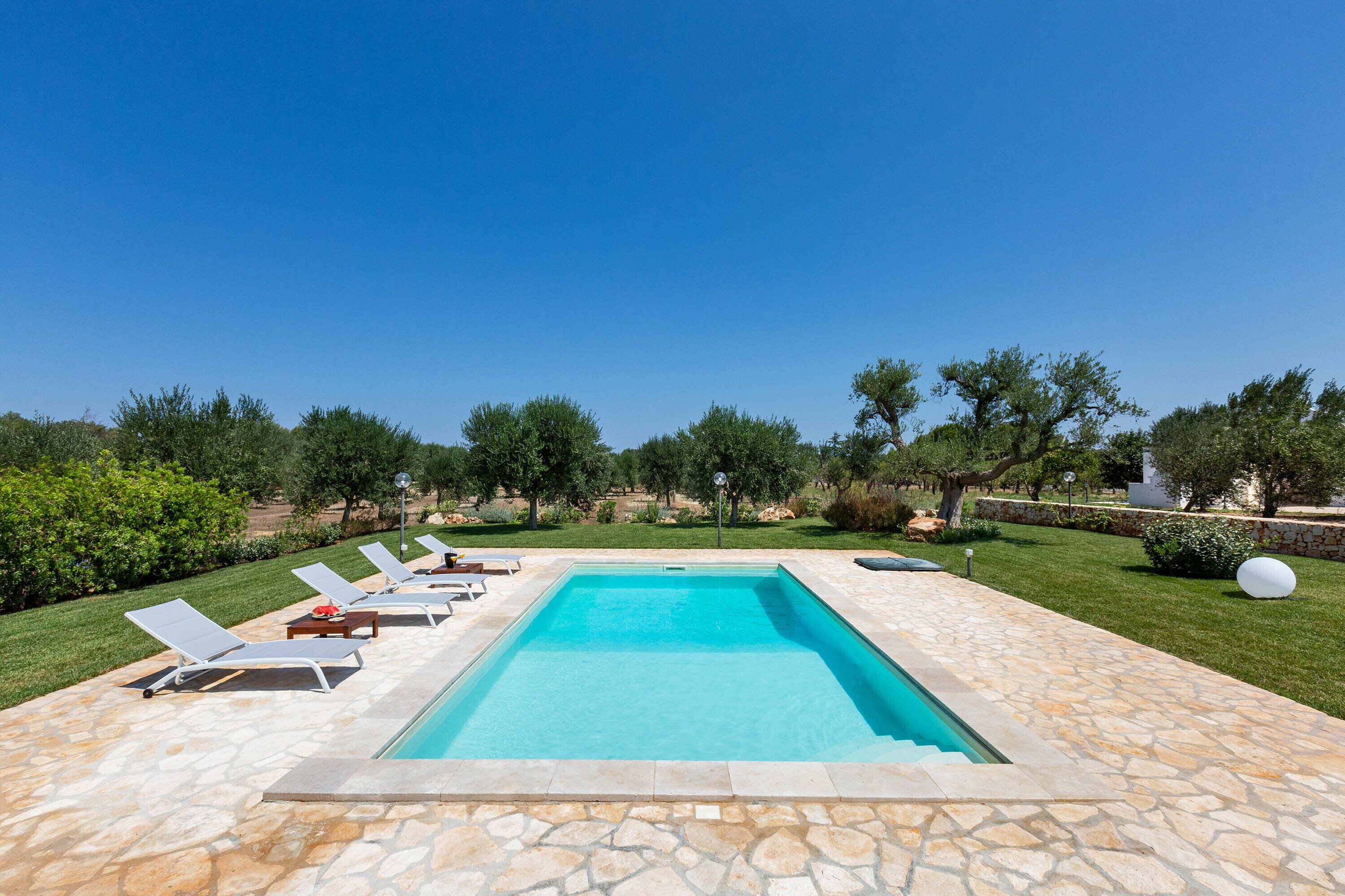 Une piscine rectangulaire bordée d’une terrasse en pierre, entourée de chaises longues et d’une pelouse bien entretenue.