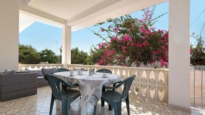 Un espace extérieur abrité, aménagé pour prendre les repas, avec une table dressée pour deux personnes, entourée de chaises et agrémenté d’un canapé.