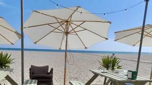 Een terras aan het strand met buitenmeubilair, parasols en uitzicht op de oceaan.