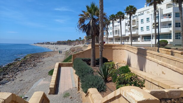 Een kustpromenade met een strand, palmbomen en moderne gebouwen.