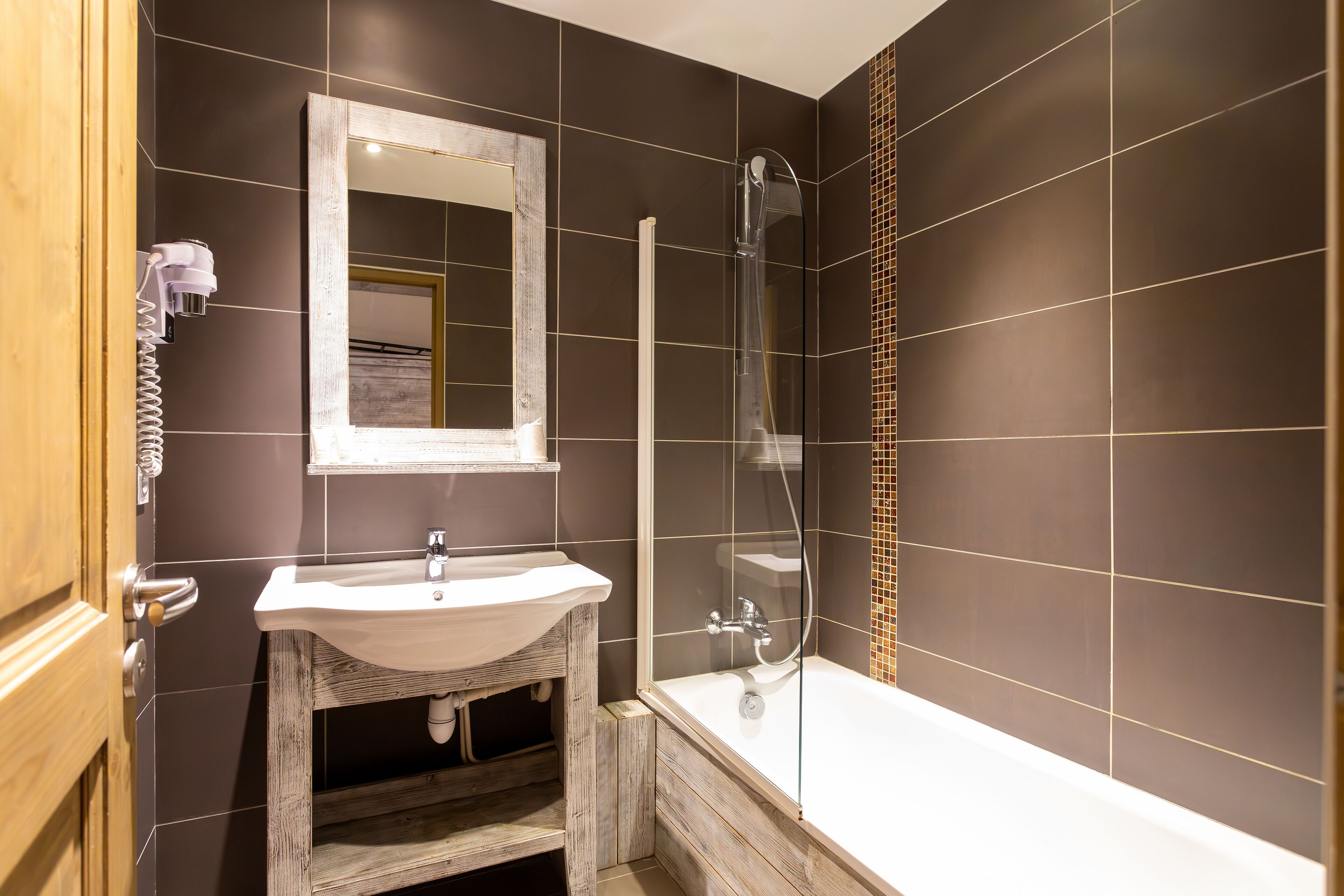 A bathroom with a wooden vanity, a white sink, a glass-enclosed shower, and dark tiled walls.