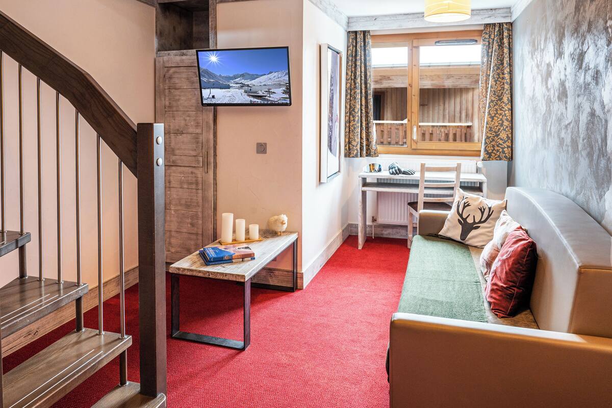 A cozy living room with a red carpet, wooden staircase, a flat-screen TV, a dining area with a table and chairs, and a sofa with cushions.