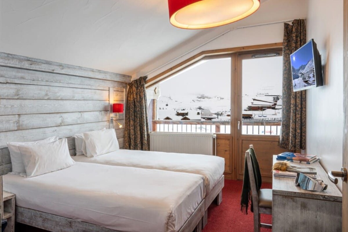 A hotel room with two beds, a red lamp, a desk with a chair, a window with a view of mountains, and a TV mounted on the wall.