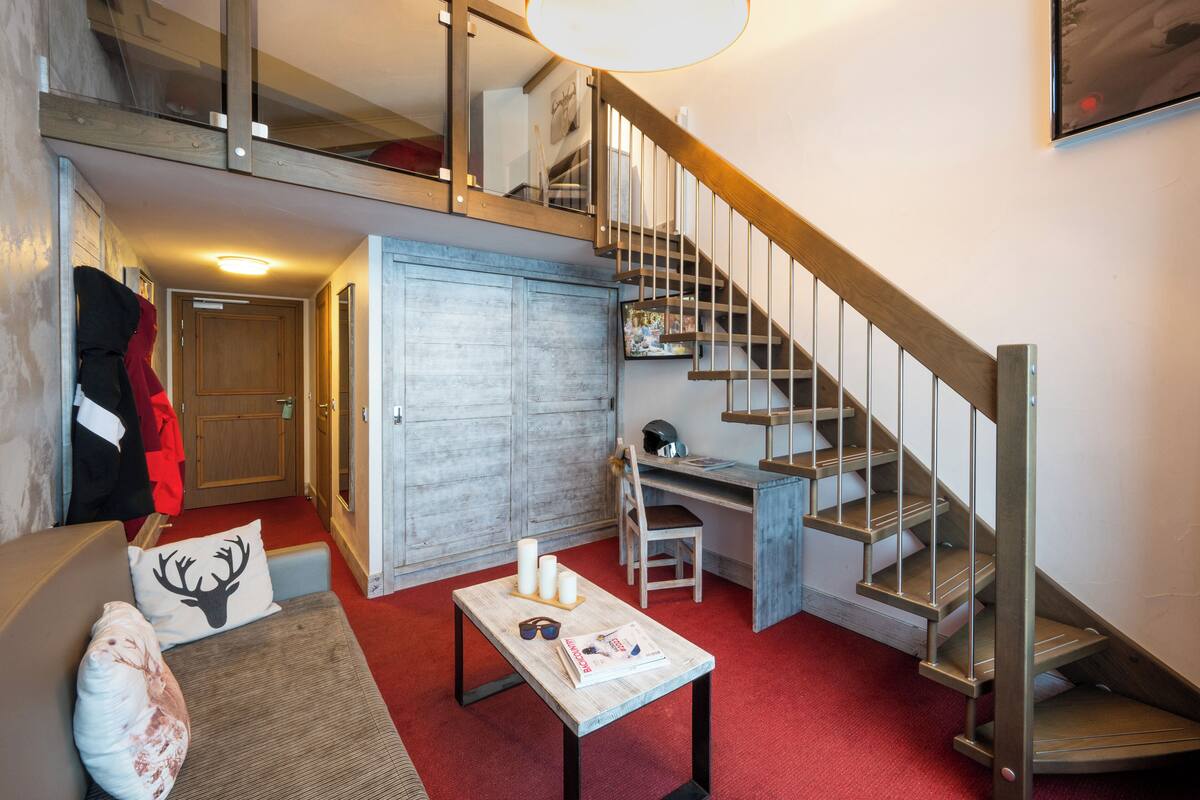 A compact living space with a loft area, a desk, a chair, a sofa, and a staircase.