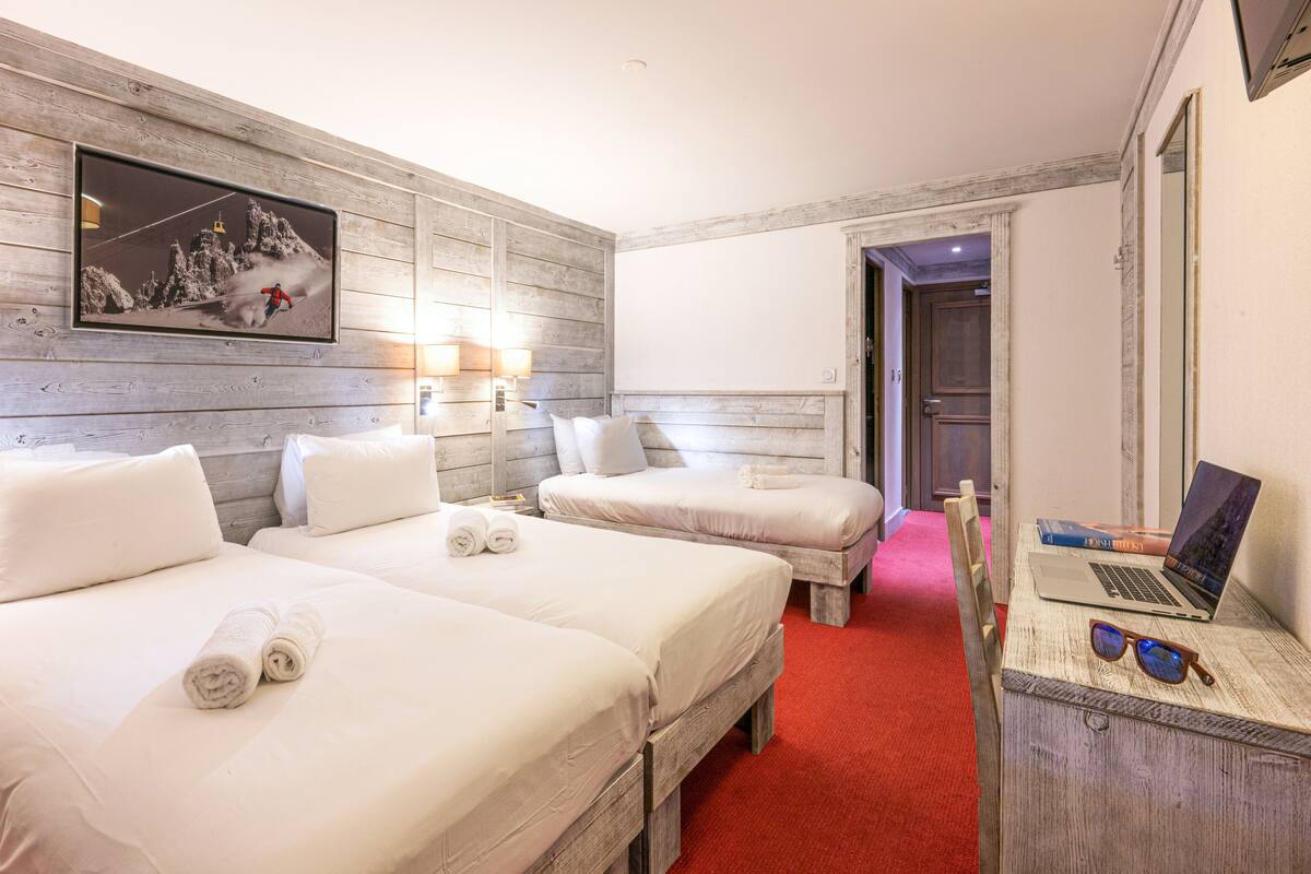 A hotel room with two beds, a TV mounted on the wall, a desk with a laptop and sunglasses, and a red carpet.