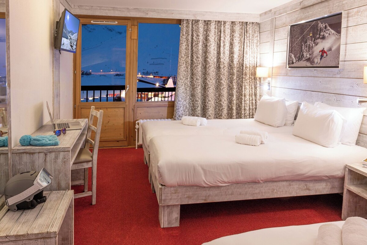 A hotel room with a large bed, a desk with a laptop, a chair, a TV, and a view of a snowy landscape through a glass door.