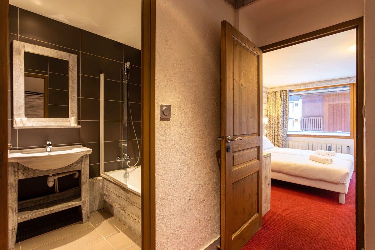 A bathroom with a sink, mirror, and shower, and a bedroom with a bed and window.