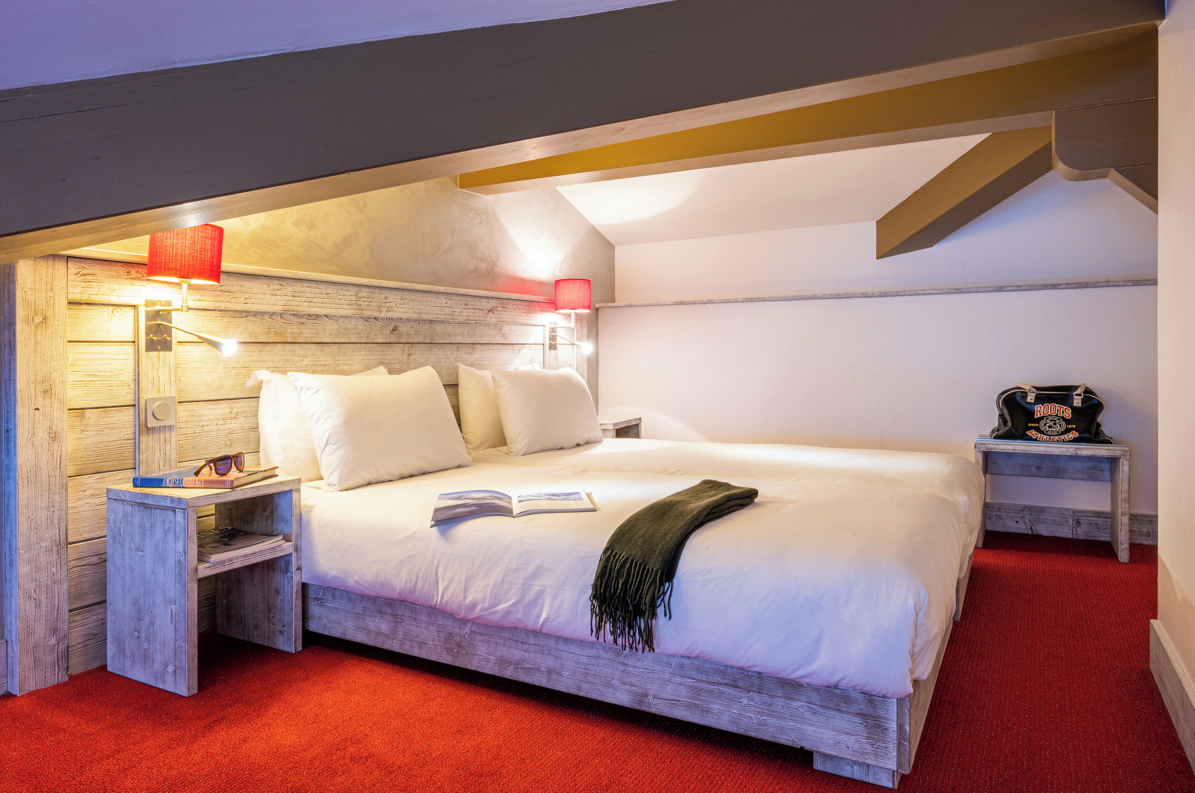 Un dormitorio con cabecera de madera, alfombra roja, cama con ropa de cama blanca, mesita de noche con lámpara y una pequeña mesa con un libro y gafas de sol.