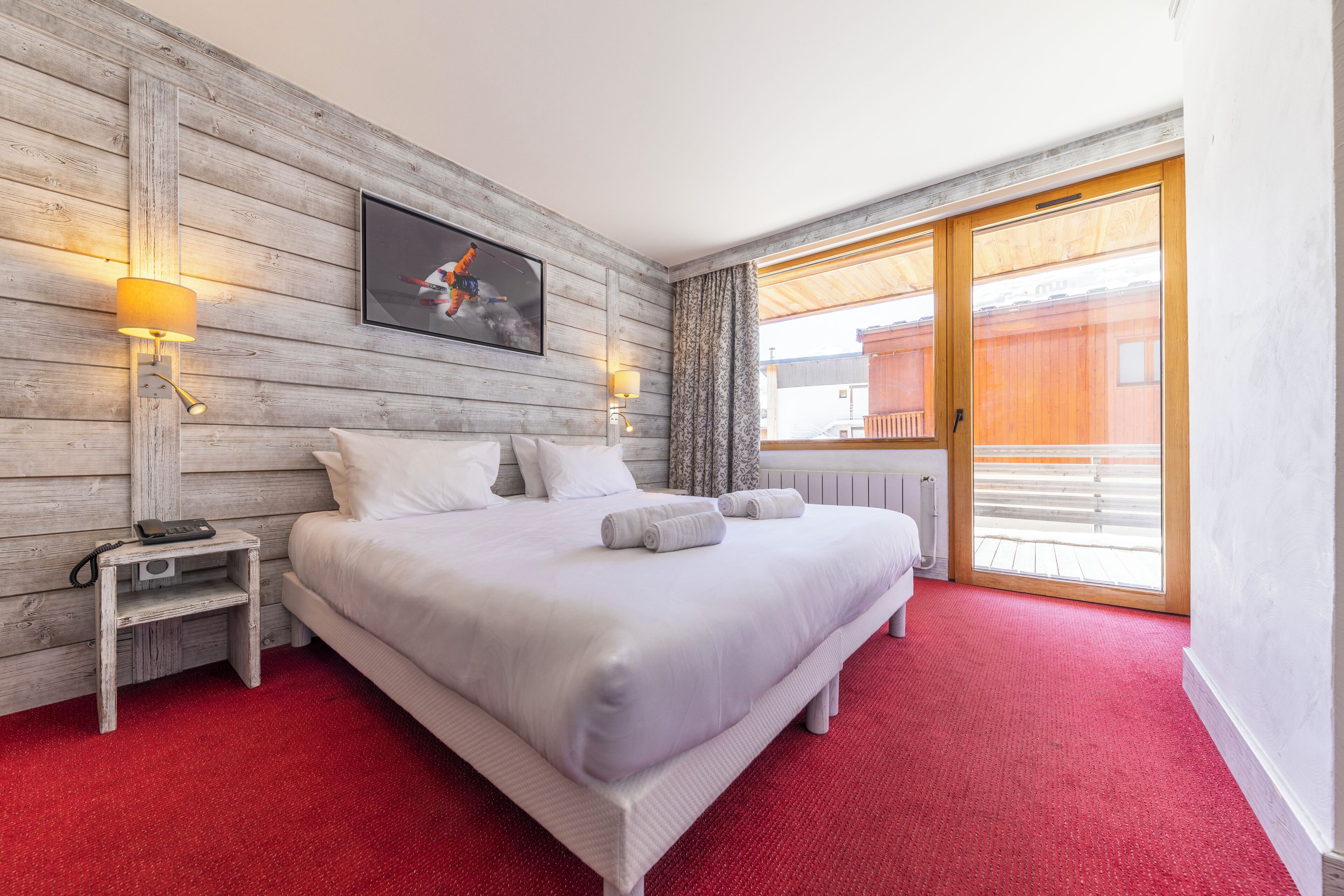 A hotel room with a wooden wall, a bed with white bedding, two bedside lamps, a small nightstand with a phone, and a large sliding glass door to the outside.