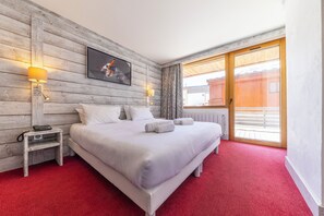 Habitación de hotel con pared de madera, cama con ropa blanca, dos lámparas de noche, una mesita de noche con teléfono y un gran ventanal corredero al exterior.