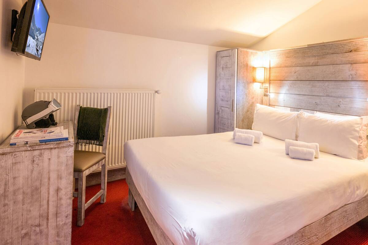 A hotel room with a wooden headboard, a bed with white linens, a desk with a chair, a radiator, and a television on the wall.
