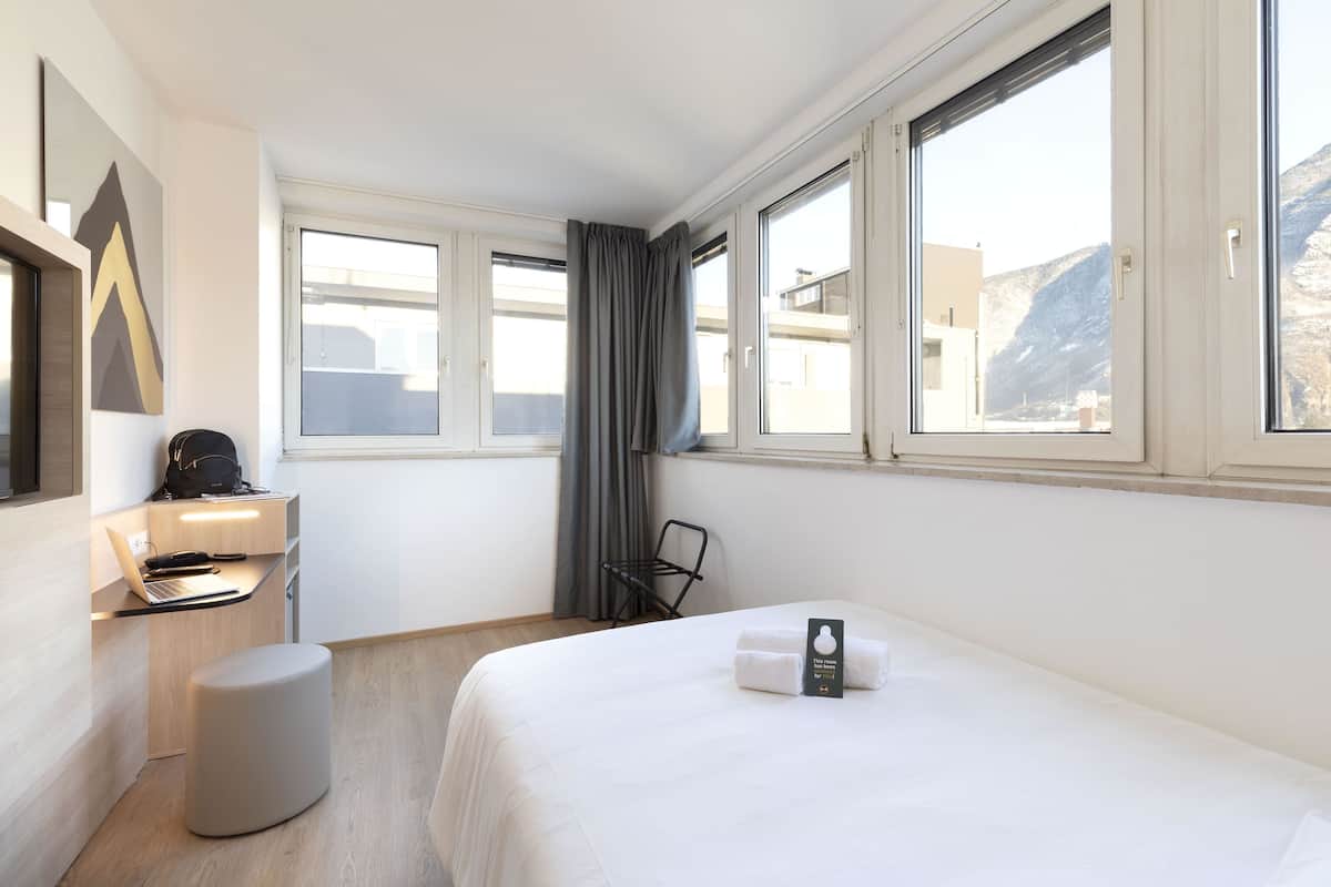 Une chambre d’hôtel avec un lit, un bureau avec un ordinateur portable, une chaise et une fenêtre avec des rideaux.