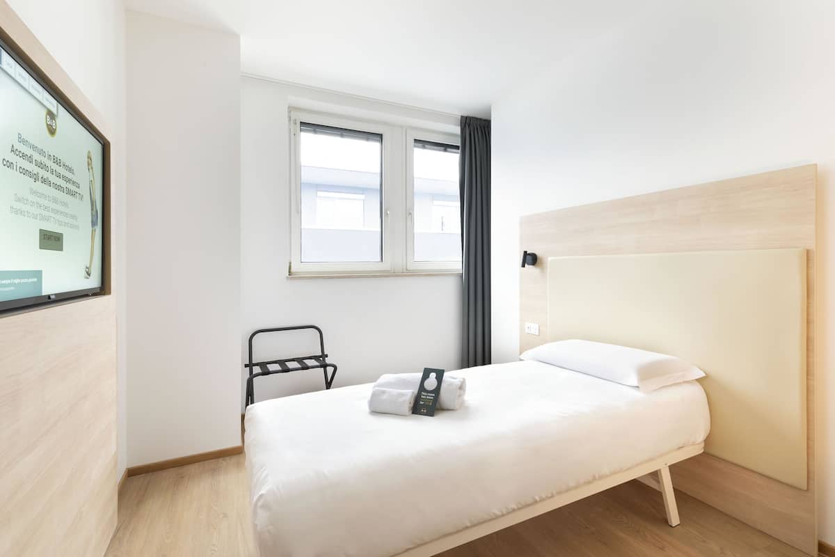 Un lit bien fait, avec une tête de lit, une fenêtre avec des rideaux et un écran fixé au mur qui affiche un message de bienvenue.