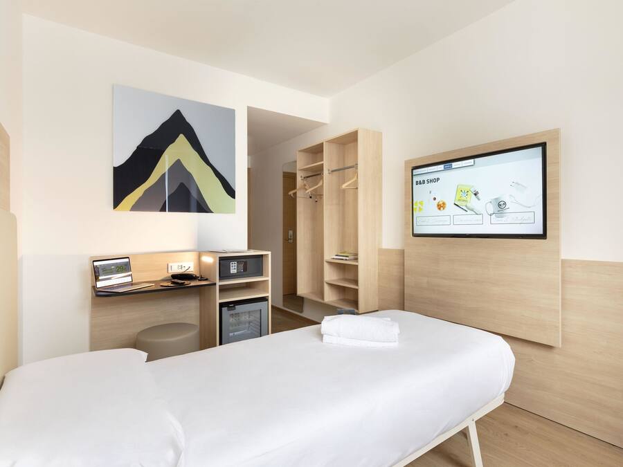 Quarto de hotel com cama, secretária com computador, televisão exibindo um aplicativo de compras e guarda-roupa.
