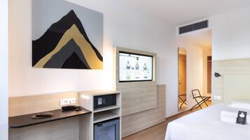 Una habitación de hotel moderna con una cama, un escritorio con ordenador, un refrigerador pequeño, un televisor montado en la pared y una obra de arte colgada en la pared.