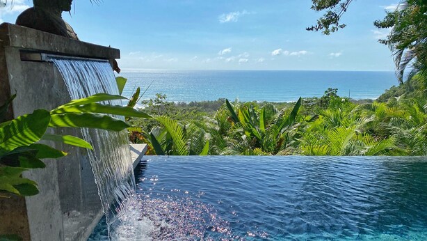 Una piscina d'hidromassatge infinita amb cascada, envoltada de vegetació exuberant i vistes a l'oceà.