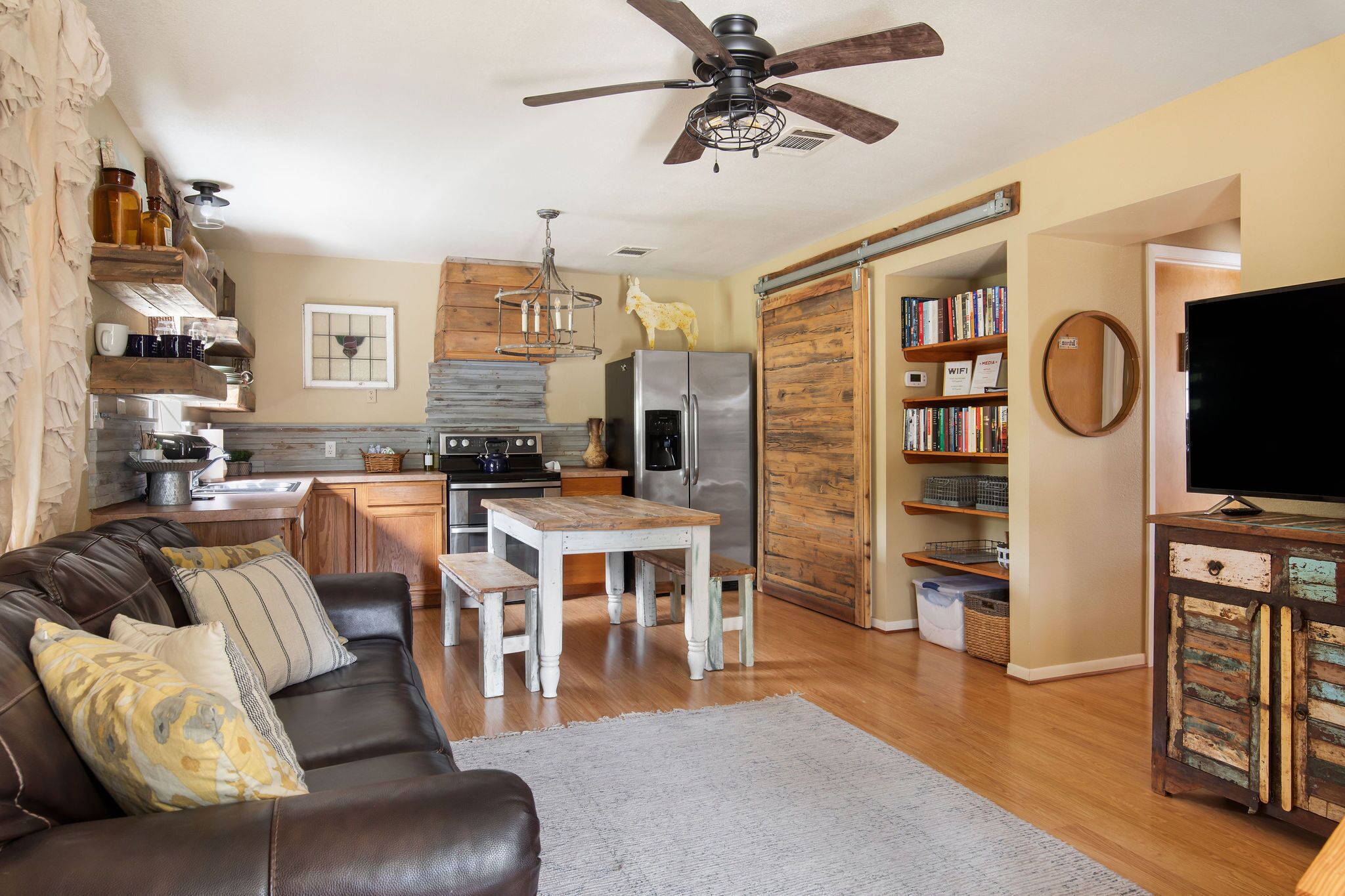 A modern kitchen with a refrigerator, stove, and wooden cabinets. There is a dining table with benches and a ceiling fan.