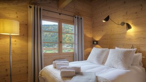 A wooden cabin bedroom with a bed, two bedside lamps, a window with curtains, and a view of a forested area. - Chalet Cosyneige for 11 in La Clusaz, hot tub - OVO Network (La Clusaz)