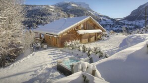 A wooden chalet with a snow-covered roof, a hot tub, and a view of a snowy mountain landscape. - Chalet Cosyneige for 11 in La Clusaz, hot tub - OVO Network (La Clusaz)