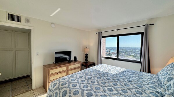 Un dormitorio con una cama, un televisor sobre una cómoda de madera, una ventana con vista a la ciudad y una puerta.