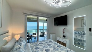 A hotel room with a bed, a TV, a balcony with a view of the sea, and a desk with a chair. - Sea Winds 705: Completely Renovated Beach Front Condo! (Marco Island)