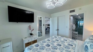 A bedroom with a bed, a dresser, a mirror, a television, and a bathroom visible through an open door. - Sea Winds 705: Completely Renovated Beach Front Condo! (Marco Island)