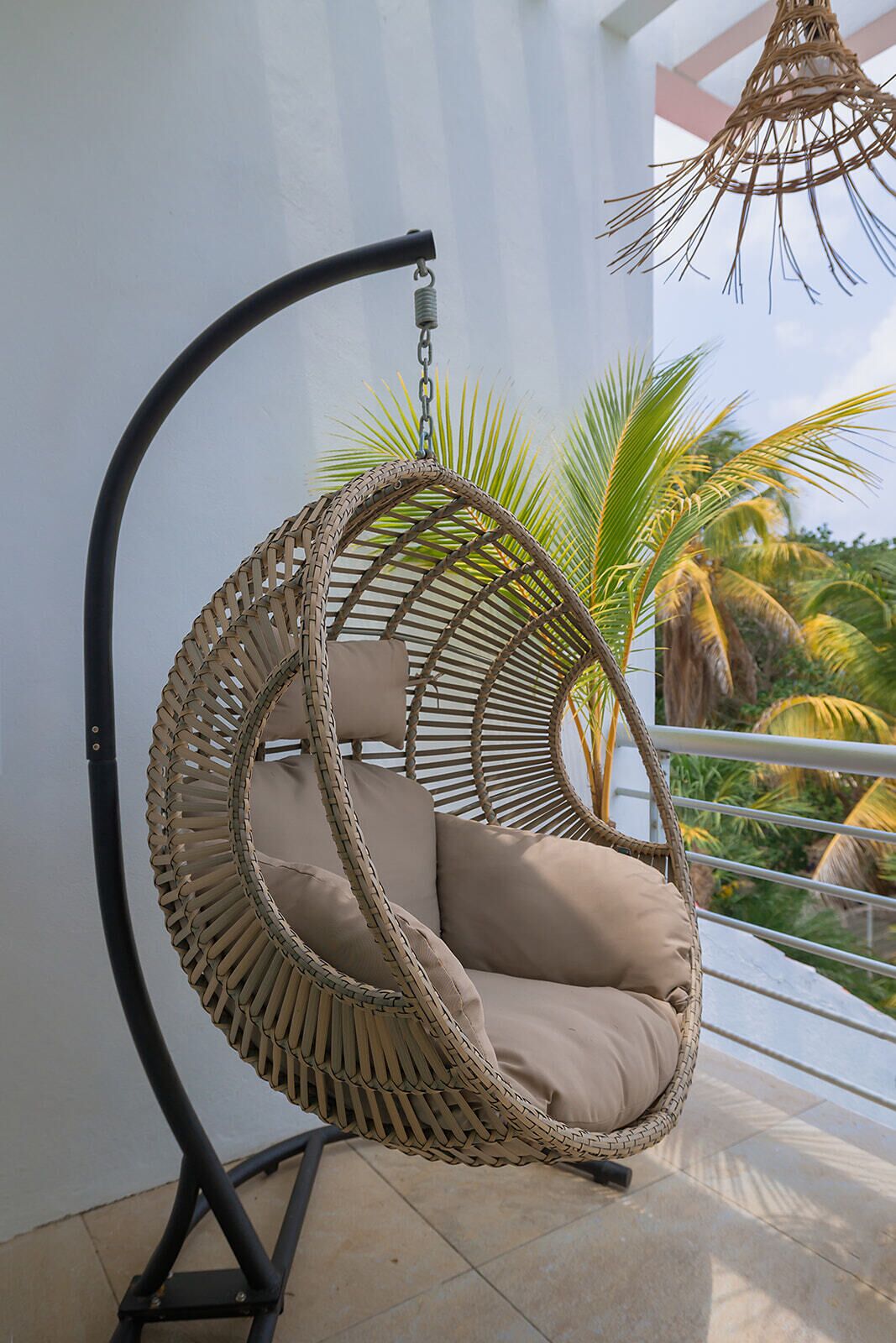 Un fauteuil suspendu en osier, agrémenté de coussins, sur un balcon offrant une vue sur des palmiers.